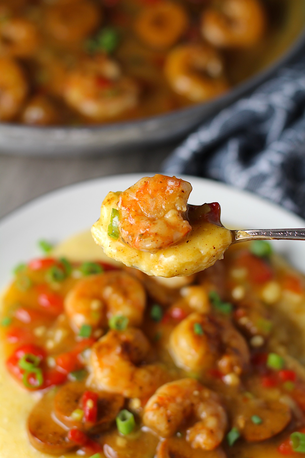 Spoon holding a bite of Cajun Shrimp and Grits recipe over a plate with pan in background. It has seared shrimp in a creamy smoky cajun sauce with chicken andouille sausage and pimiento. Served over creamy, cheesy grits.