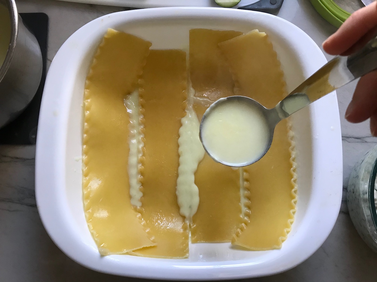 Ladle pouring white sauce over lasagna noodles in dish for White Lasagna Recipe with Zucchini and Ground Beef