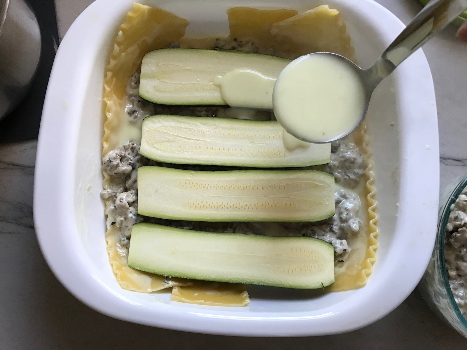 Ladle pouring white sauce over zucchini slices on top of beef and lasagna noodles in dish for White Lasagna Recipe with Zucchini and Ground Beef