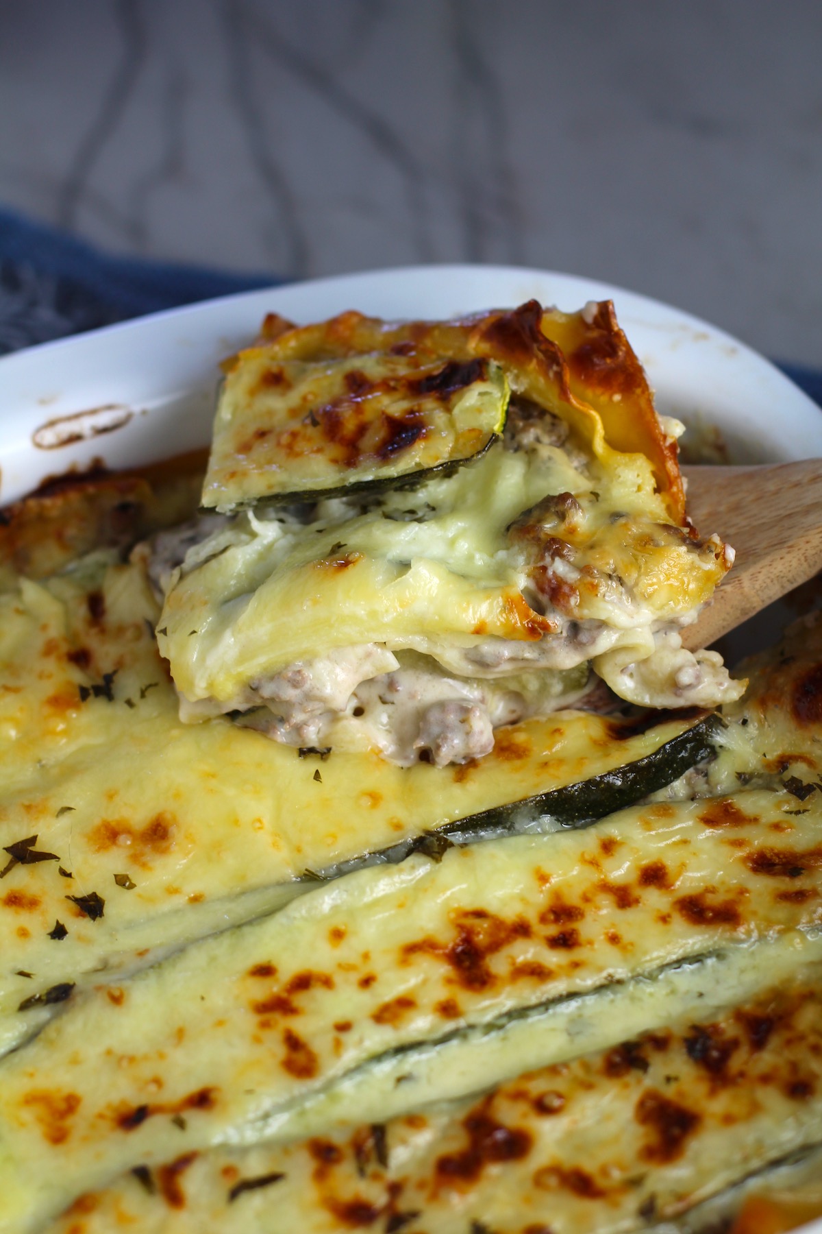 Spatula lifting a piece of White Lasagna Recipe with Zucchini and Ground Beef with browned melted cheese on top in casserole dish! It's a make-ahead family dinner! It's creamy, hearty, and delicious with layers of noodles, zucchini, ground beef, creamy cheese, and a silky white sauce. 