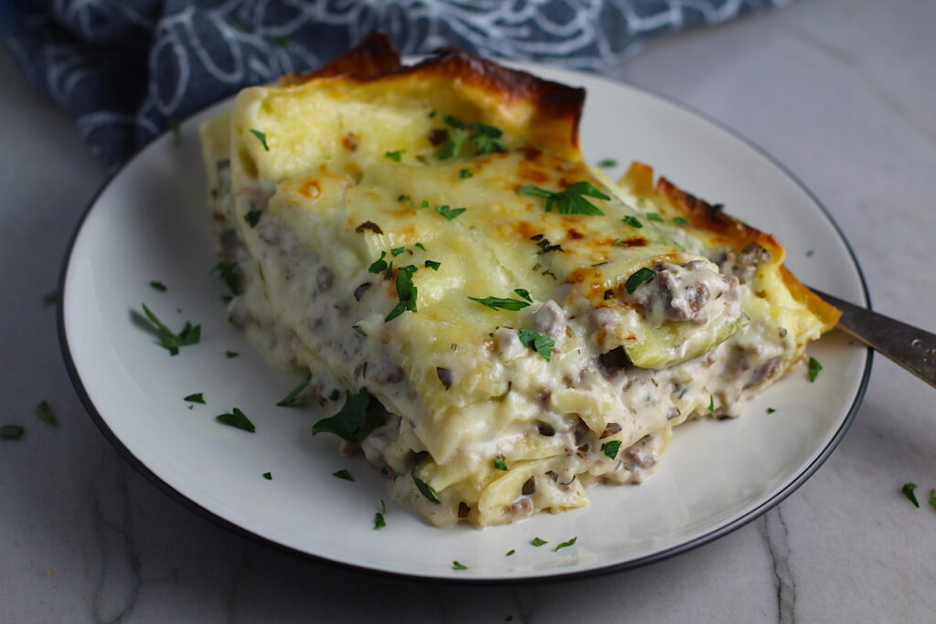 Piece of White Lasagna Recipe with Zucchini and Ground Beef with browned melted cheese on top on a plate with parsley. It's a make-ahead family dinner! It's creamy, hearty, and delicious with layers of noodles, zucchini, ground beef, creamy cheese, and a silky white sauce.