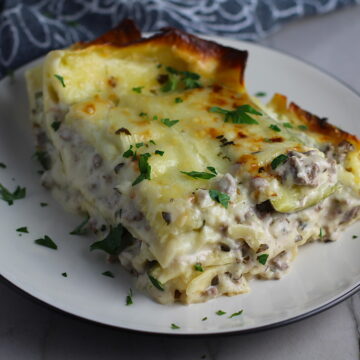 Piece of White Lasagna Recipe with Zucchini and Ground Beef with browned melted cheese on top on a plate with parsley. It's a make-ahead family dinner! It's creamy, hearty, and delicious with layers of noodles, zucchini, ground beef, creamy cheese, and a silky white sauce.