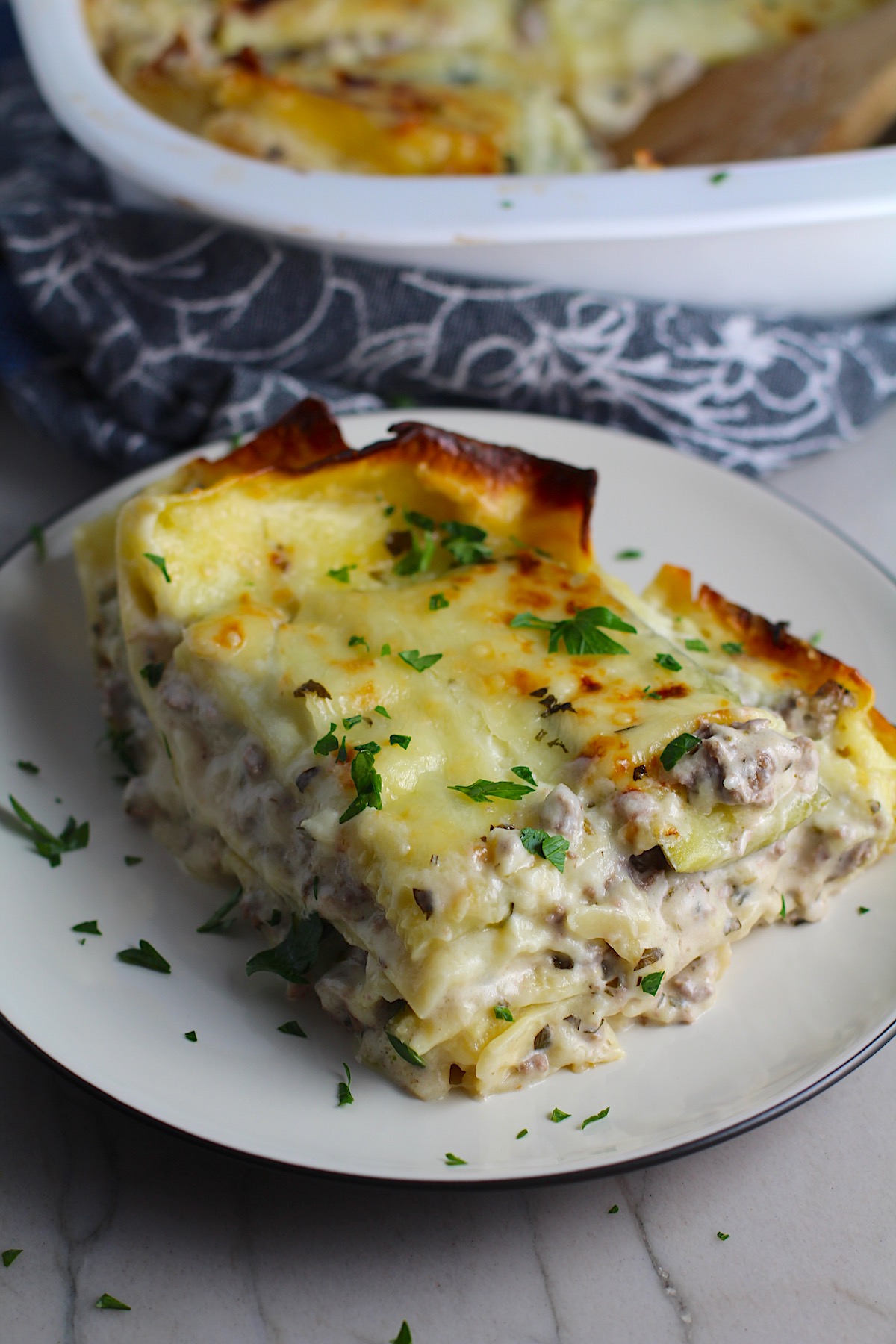Piece of White Lasagna Recipe with Zucchini and Ground Beef with browned melted cheese on top on a plate with parsley. It's a make-ahead family dinner! It's creamy, hearty, and delicious with layers of noodles, zucchini, ground beef, creamy cheese, and a silky white sauce.