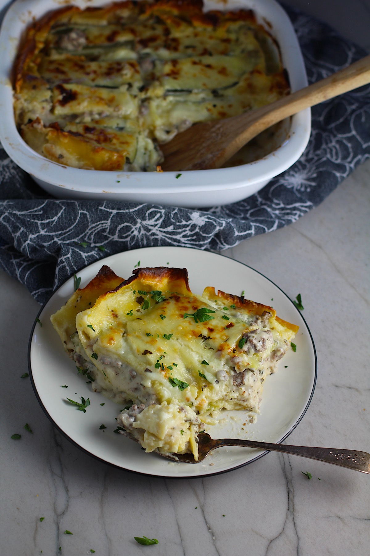 Fork with a piece of White Beef Lasagna with Cream Cheese and Zucchini with browned melted cheese on top on a plate with parsley. It's a make-ahead family dinner! It's creamy, hearty, and delicious with layers of noodles, zucchini, ground beef, creamy cheese, and a silky white sauce.
