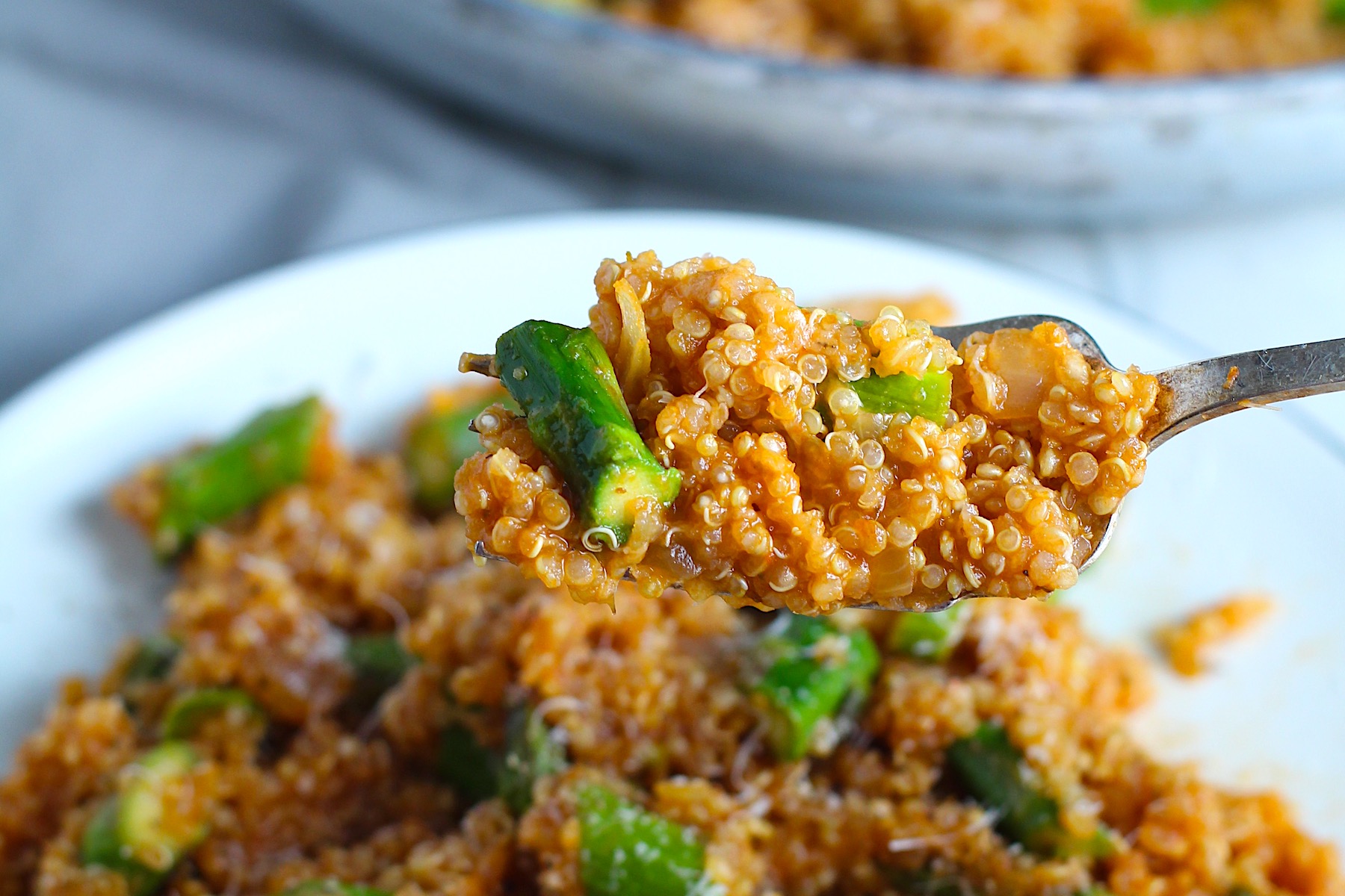 Fork holding a bite of Asparagus and Tomato Quinoa Risotto with parmesan over a plate of food with pan in background.