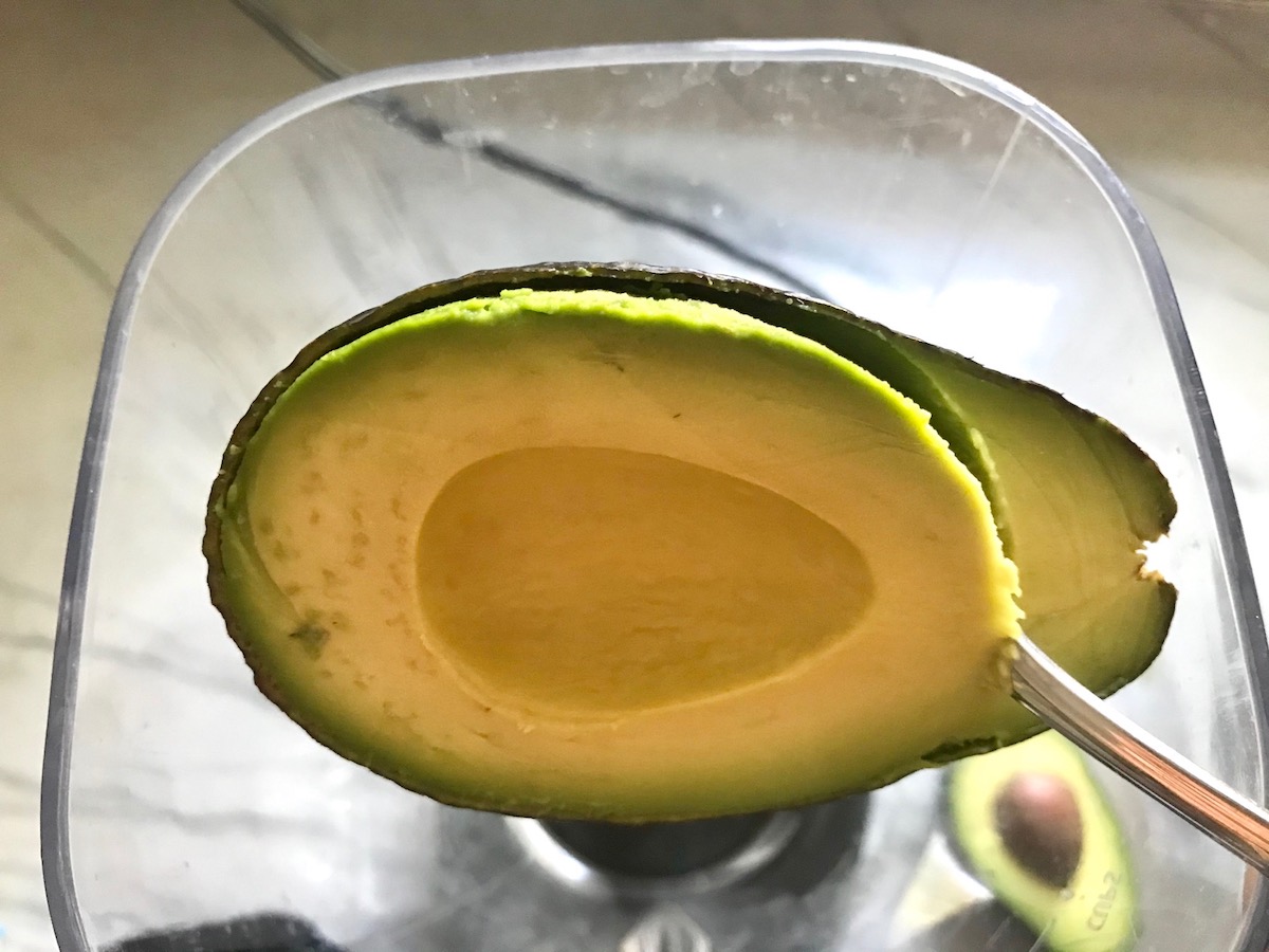 Spoon scooping avocado out of skin into a blender for Avocado Crema Recipe. It goes on everything! It's a creamy, tangy, rich and delicious sauce that's made in just minutes in the blender!