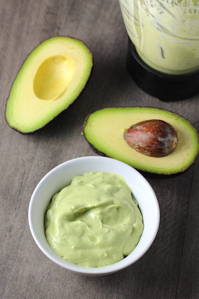 Easy Avocado Crema Recipe in a bowl on table with a cut open avocado with pit and blender in back. It goes on everything! It's a creamy, tangy, rich and delicious sauce that's made in just minutes in the blender!