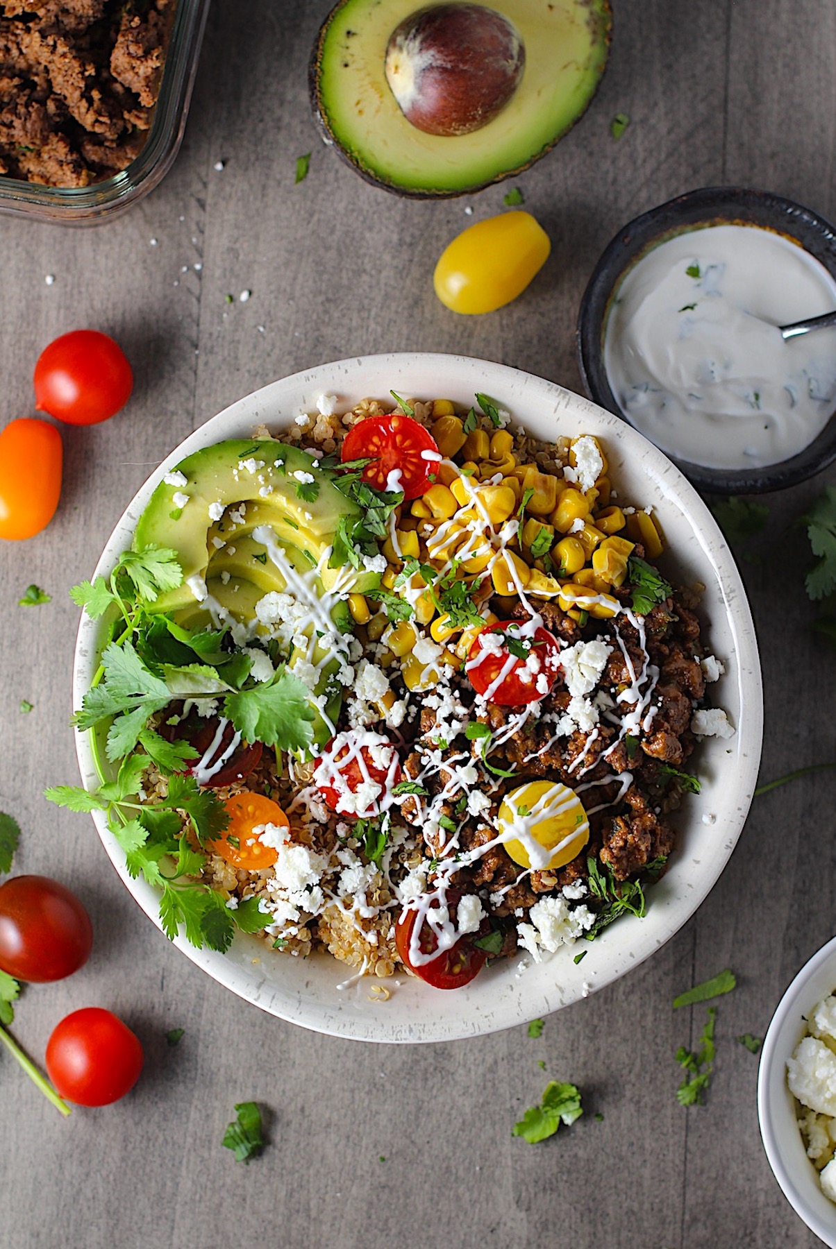 Taco Quinoa Bowl with Ground Beef, corn, cotija cheese, tomatoes, fresh cilantro, and cilantro lime crema drizzled on top. Ground beef bowl, avocado half with pit, bowl of crema, tomatoes and cilantro on the table.