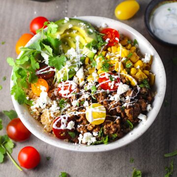 Taco Quinoa Bowl with Ground Beef, corn, cotija cheese, tomatoes, fresh cilantro, and cilantro lime crema drizzled on top. Bowl of crema, bowl of cotija cheese, tomatoes and cilantro on the table.