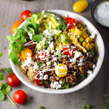Taco Quinoa Bowl with Ground Beef, corn, cotija cheese, tomatoes, fresh cilantro, and cilantro lime crema drizzled on top. Bowl of crema, bowl of cotija cheese, tomatoes and cilantro on the table.