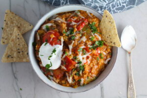 Bowl of Tex Mex Unstuffed Peppers with Ground Chicken with a dollop of sour cream and a tortilla chip dipped in. Juicy Sweet Bell Peppers are chopped up and cooked with the Tex Mex style ground chicken, tomatoes, Mexican seasonings, cheese, rice and corn tortillas.