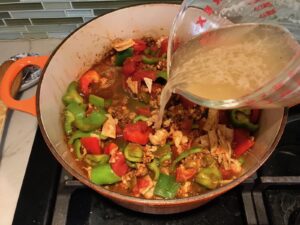Pouring broth into pot with ground chicken cooking, peppers, and tomatoes for Tex Mex Unstuffed Peppers with Ground Chicken. Juicy Sweet Bell Peppers are chopped up and cooked with the Tex Mex style ground chicken, tomatoes, Mexican seasonings, cheese, rice and corn tortillas.