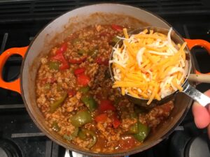 Adding shredded cheese to pot of cooked Tex Mex Unstuffed Peppers with Ground Chicken. Juicy Sweet Bell Peppers are chopped up and cooked with the Tex Mex style ground chicken, tomatoes, Mexican seasonings, cheese, rice and corn tortillas.