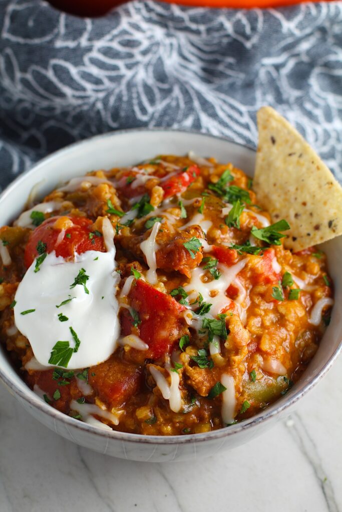 Bowl of Tex Mex Unstuffed Peppers with Ground Chicken with a dollop of sour cream and a tortilla chip dipped in. Juicy Sweet Bell Peppers are chopped up and cooked with the Tex Mex style ground chicken, tomatoes, Mexican seasonings, cheese, rice and corn tortillas.