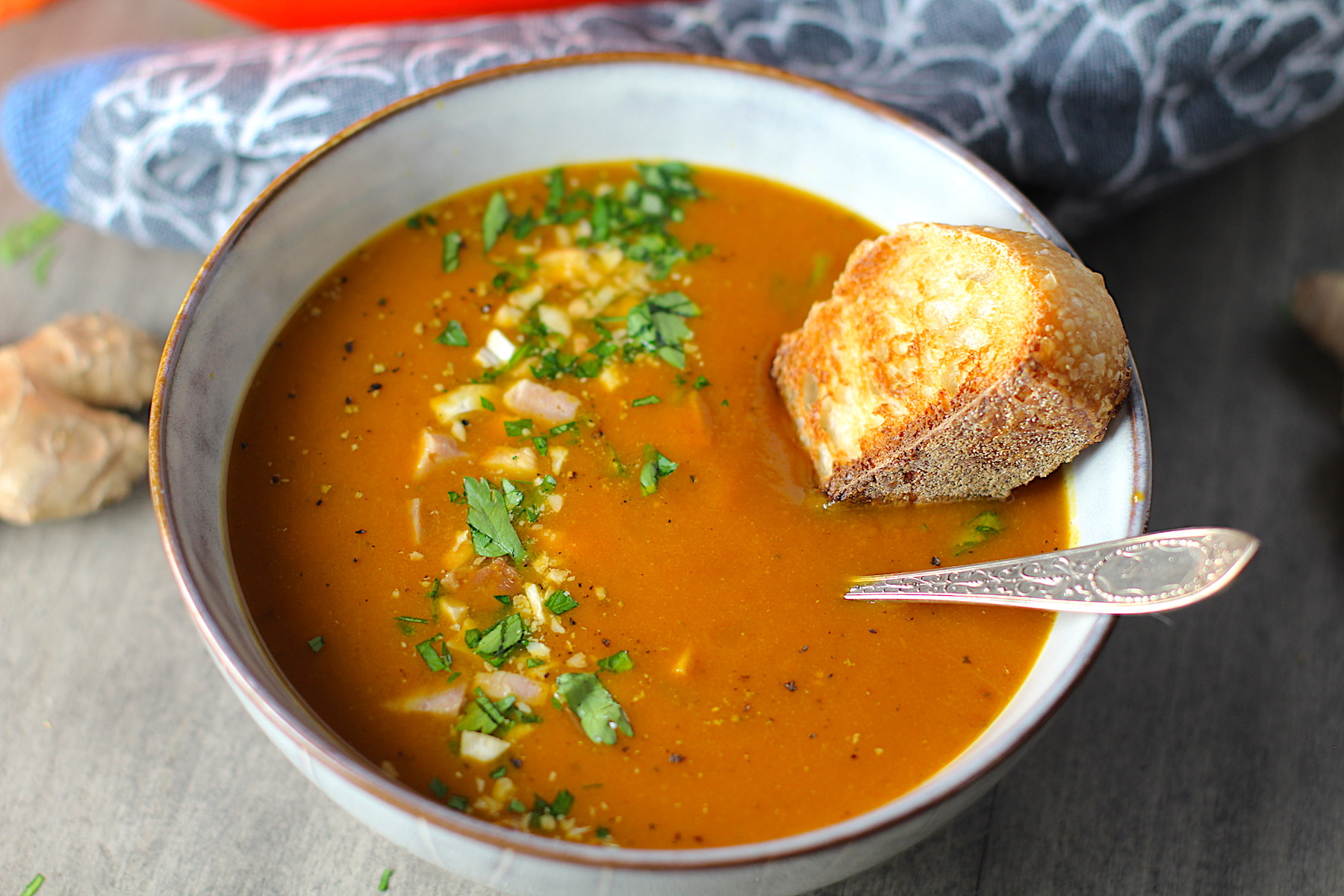Golden Carrot Ginger Soup Recipe in a bowl with spoon and bread dipped in side and parsley, chopped cashews, and ham garnish on top. Ginger root and carrot pieces on table