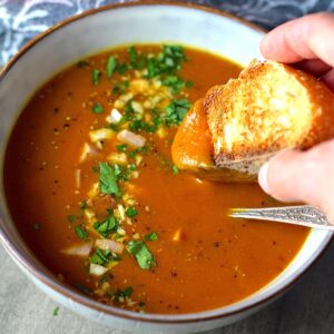 Golden Carrot Ginger Soup Recipe in a bowl with spoon and hand dipping bread in and parsley, chopped cashews, and ham garnish on top.