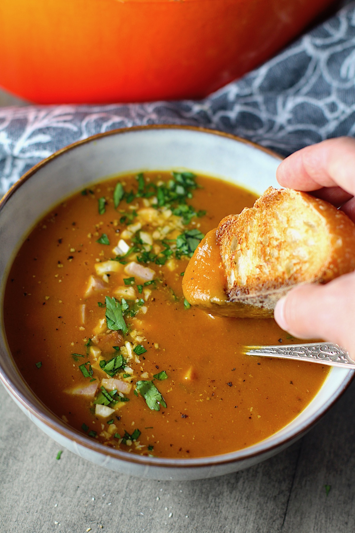 Golden Carrot Ginger Soup Recipe in a bowl with spoon and bread dipped in side and parsley, chopped cashews, and ham garnish on top. Ginger root and carrot pieces on table