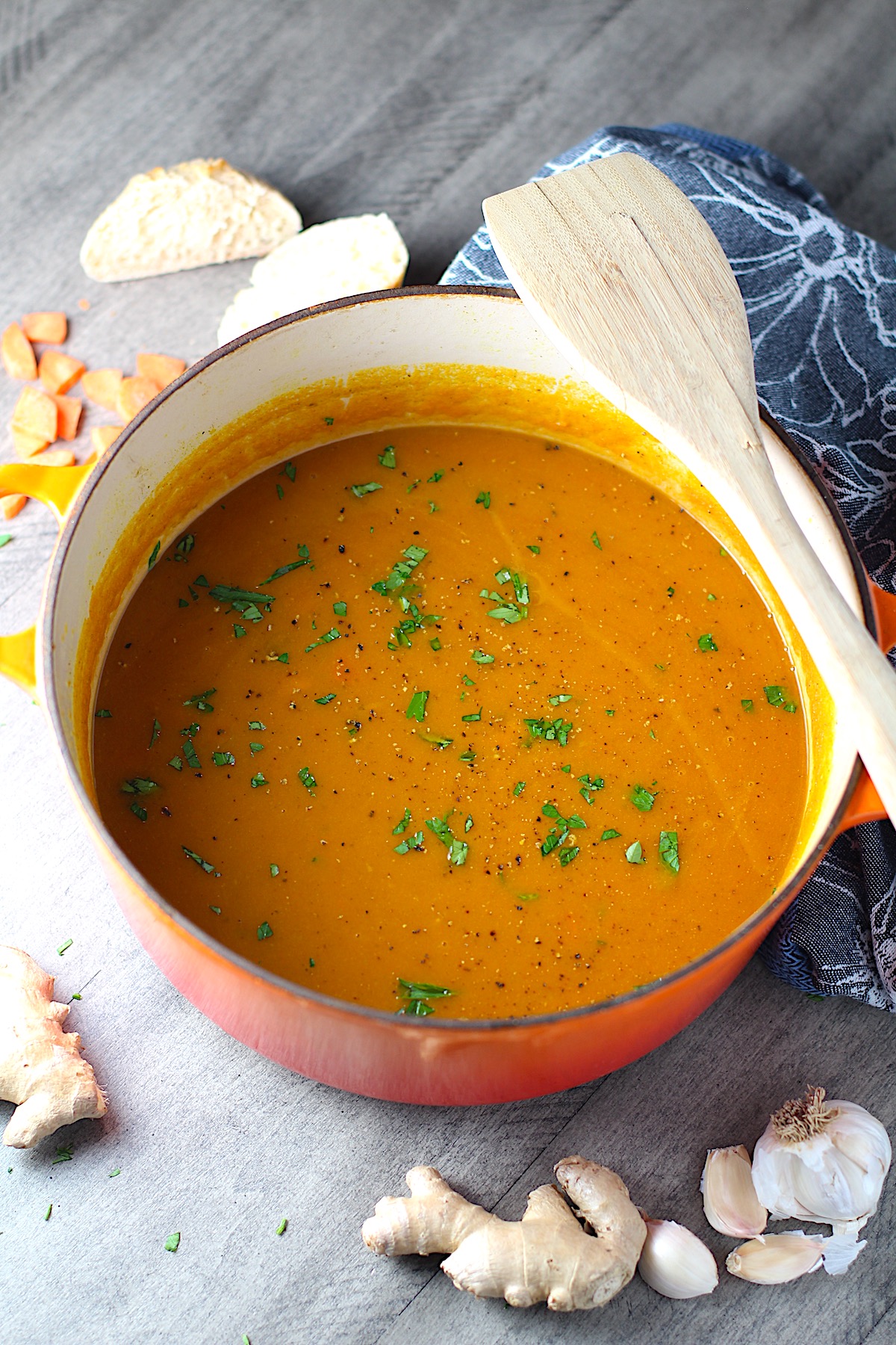 Golden Carrot Ginger Soup Recipe in a pot with wood spoon on side and parsley garnish on top. Ginger root and carrot pieces on table