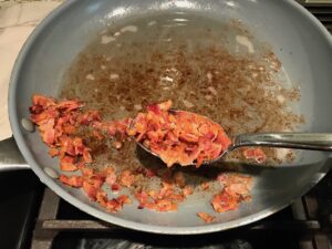 Spoon scooping out cooked diced bacon in large frying pan for Velvety Barley Recipe with Bacon and Gruyere Cheese. It's creamy, rich, nutty, smokey, hearty, and utterly delicious.