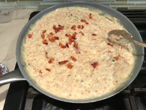 Spoon mixing cooked bacon bits into Creamy Barley Recipe with Bacon and Gruyere Cheese in large pan on stove. It's creamy, rich, nutty, smokey, hearty, and utterly delicious.