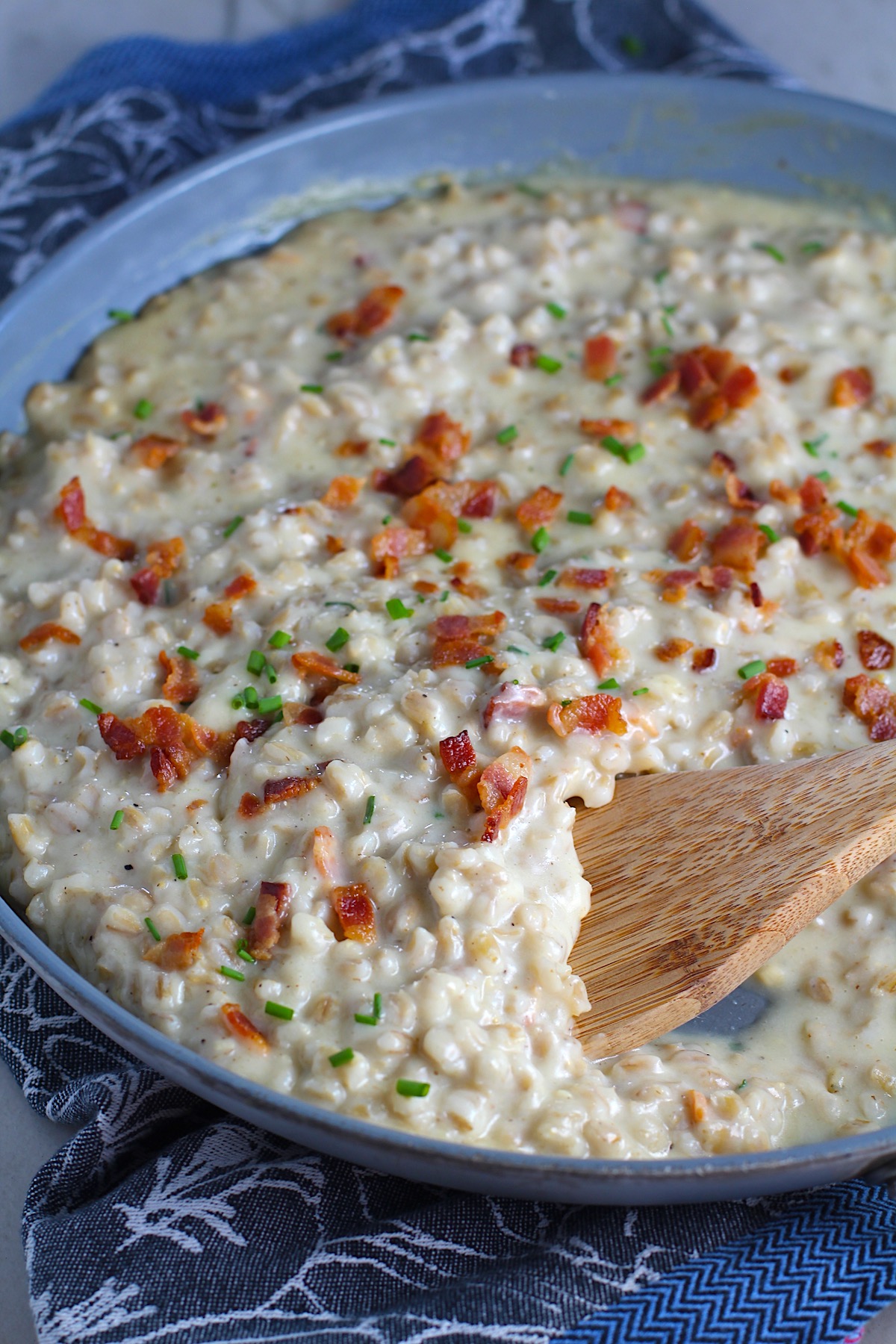Creamy Barley Recipe with Bacon and Gruyere Cheese in pan with wood spoon and crispy bacon bits and diced chives on top. It's creamy, rich, nutty, smokey, hearty, and utterly delicious.