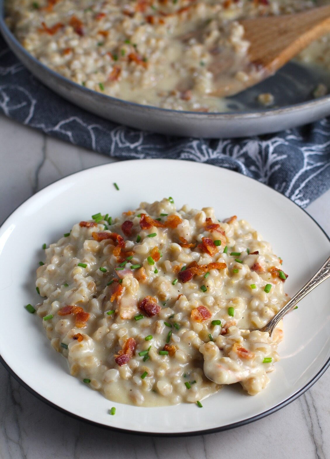 Creamy Barley Recipe with Bacon and Gruyere Cheese on a plate with pan in back and crispy bacon bits and diced chives on top and spoon with scoop on side of plate. It's creamy, rich, nutty, smokey, hearty, and utterly delicious.