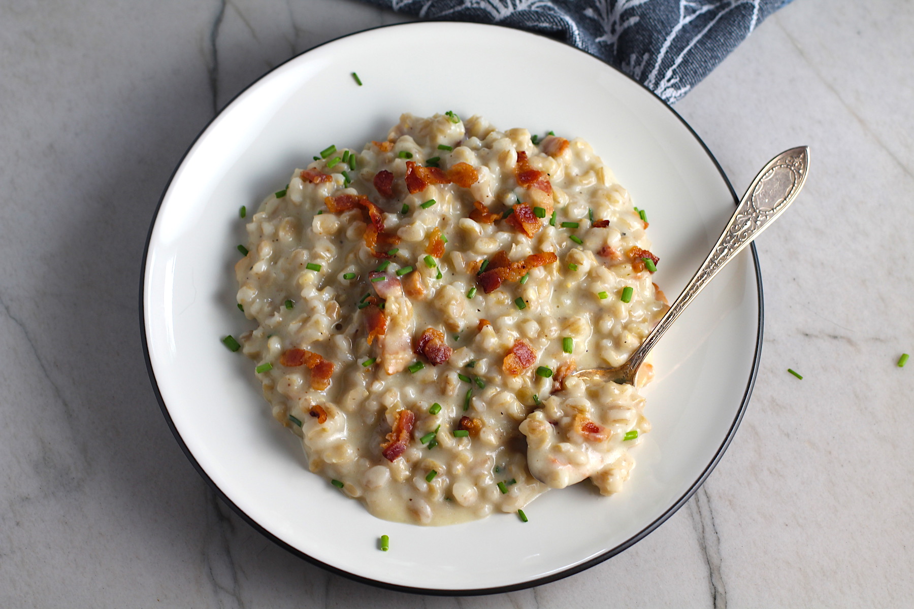 Creamy Barley with Bacon and Gruyere Cheese on plate with crispy bacon bits and diced chives on top and spoon with scoop on side of plate. It's creamy, rich, nutty, smokey, hearty, and utterly delicious.