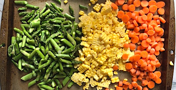 Raw cut asparagus, corn kernels, and sliced carrots on a sheet pan for Creamy Pasta Primavera. It has fettuccine, parmesan cheese, roasted asparagus, carrots, tomatoes and fresh corn kernels.