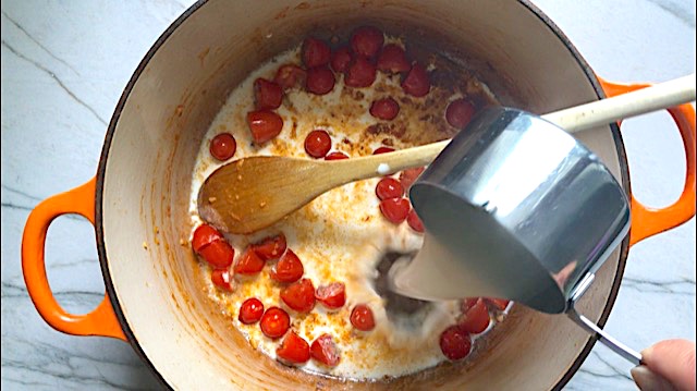 Halved grape tomatoes, half and half, and pasta water pouring in a pot for Creamy Pasta Primavera. It has fettuccine, parmesan cheese, roasted asparagus, carrots, tomatoes and fresh corn kernels.