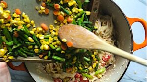 Wood spoon scraping asparagus, corn, and carrots from a sheet pan into the pot of Creamy Pasta Primavera. It has fettuccine, parmesan cheese, roasted asparagus, carrots, tomatoes and fresh corn kernels.