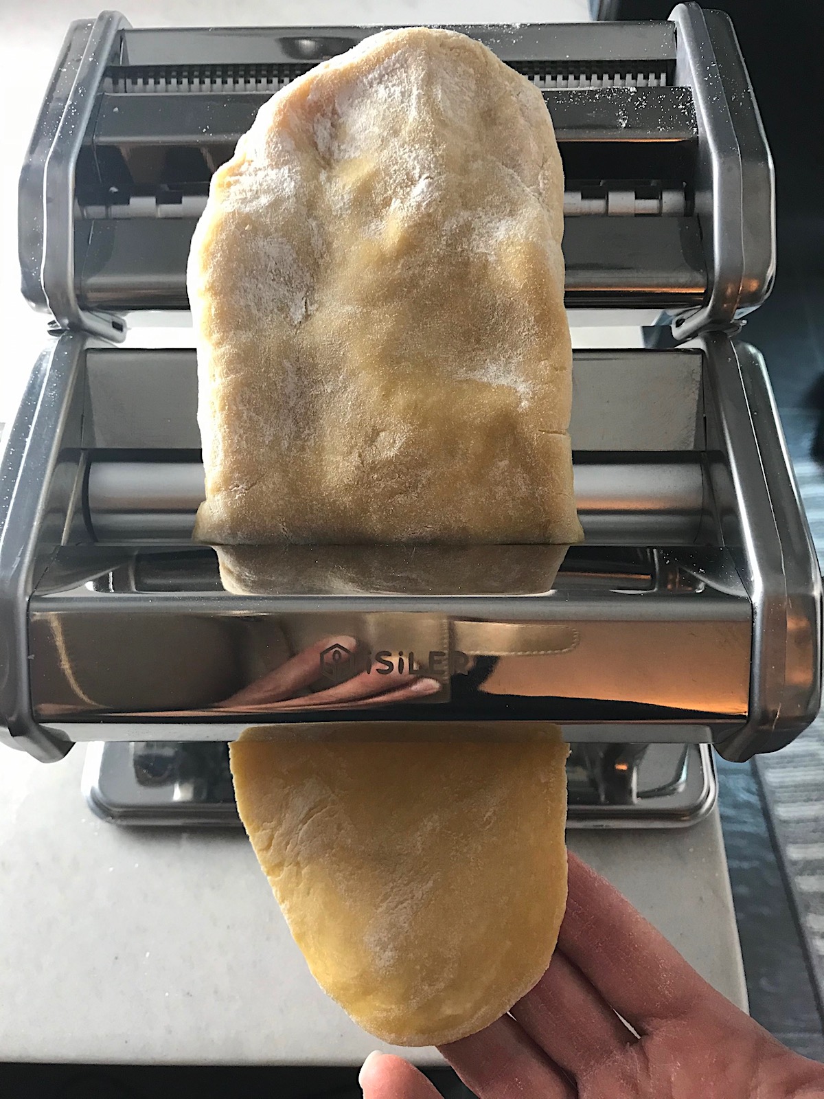 Thick piece of pasta dough going through the pasta roller with hand catching the dough at the bottom for Easy Homemade Pasta recipe.