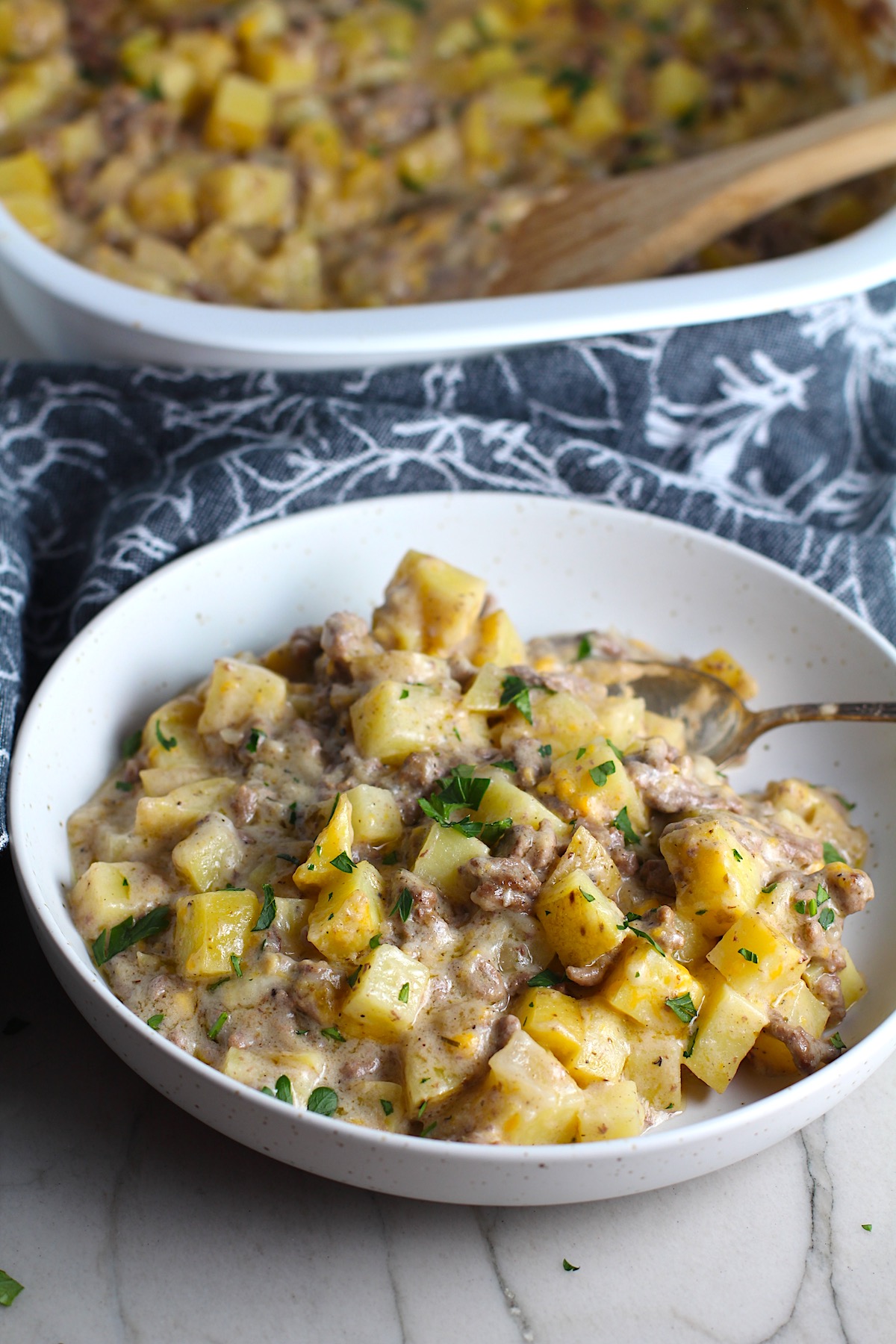 Cheesy Hamburger Potato Casserole in a bowl with a spoon and casserole in background. It's an easy, yummy, and cozy weeknight family dinner.