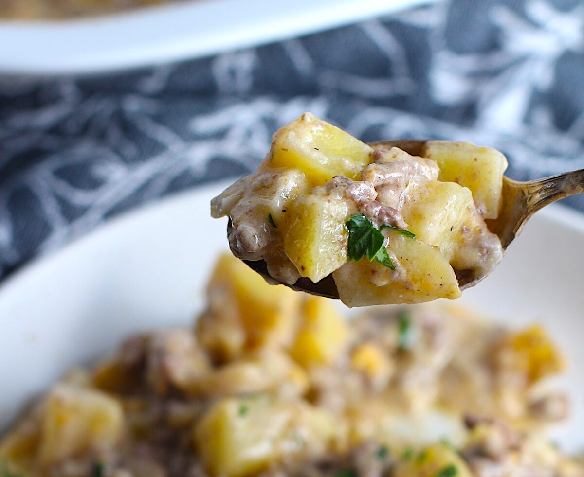 Spoon with bite of Cheesy Hamburger Potato Casserole held over a bowl. It's an easy, yummy, and cozy weeknight family dinner.