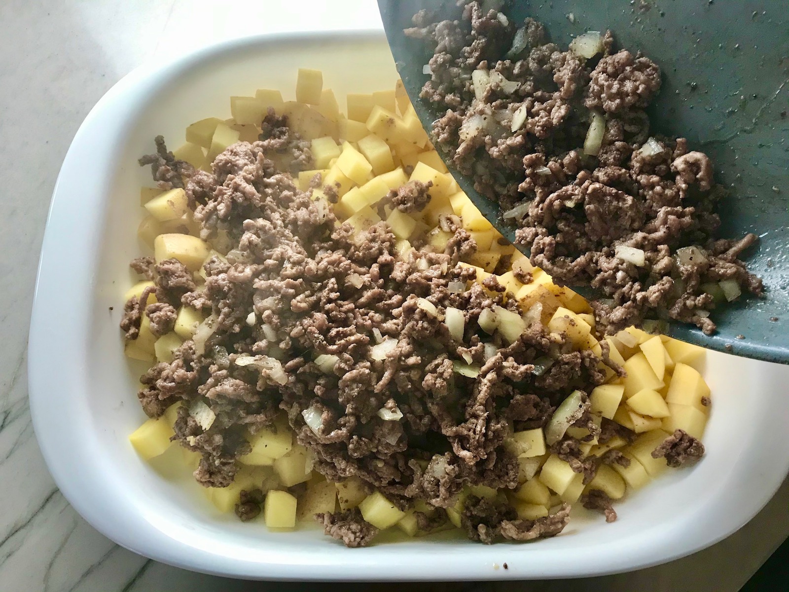 Pan pouring cooked ground beef and onions over diced raw potatoes for Cheesy Hamburger Potato Casserole. It's an easy, yummy, and cozy weeknight family dinner.