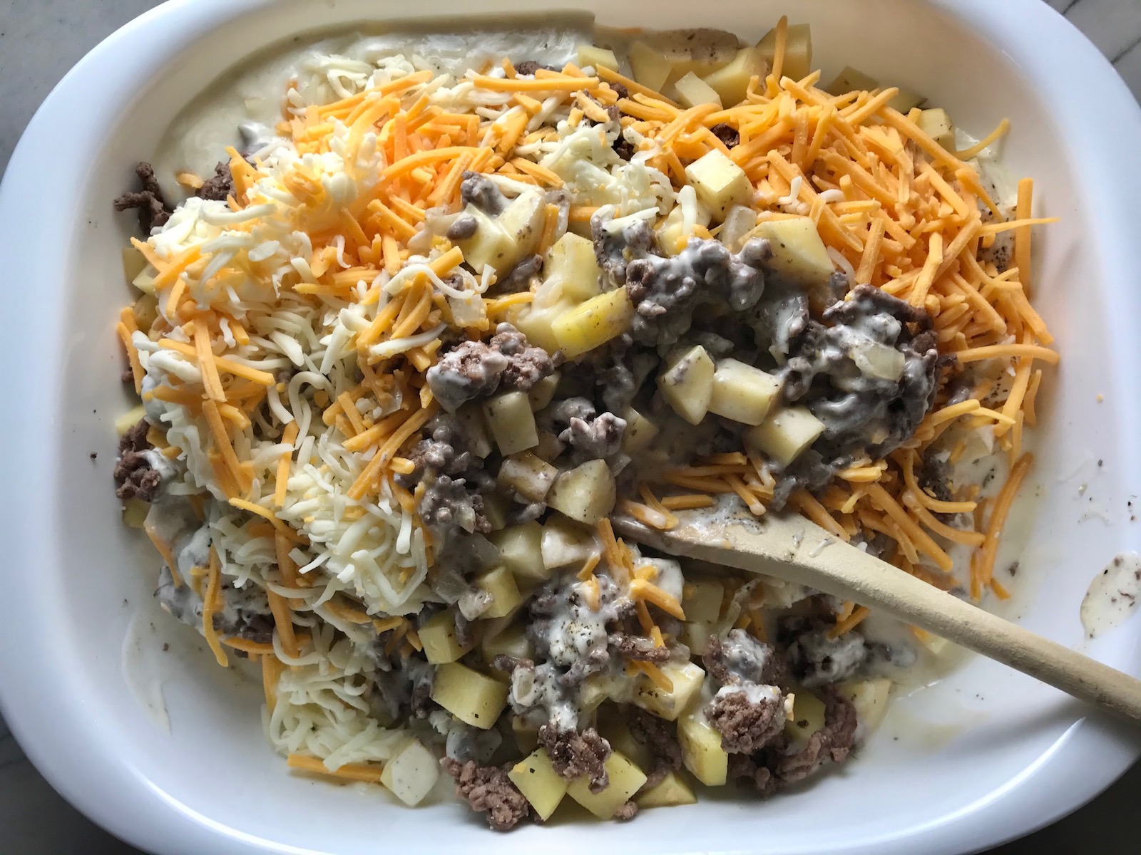 Wood spoon mixing cooked ground beef, sauce, shredded cheese, and cubed potatoes for Cheesy Hamburger Potato Casserole. It's an easy, yummy, and cozy weeknight family dinner.