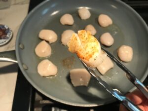 Tongs holding a scallop over pan seared side up for Glazed Pan Seared Scallops with Garlic and Honey.