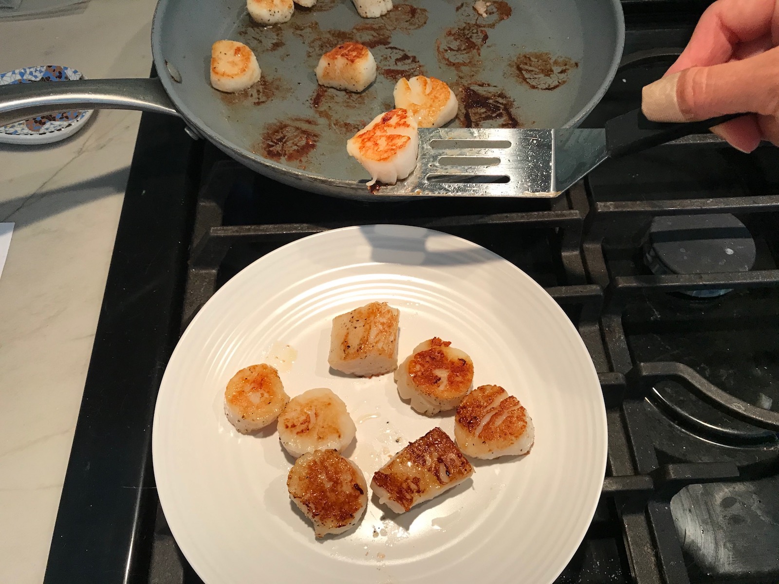 Seared scallops being transferred by spatula from pan to plate for Glazed Pan Seared Scallops with Garlic and Honey.