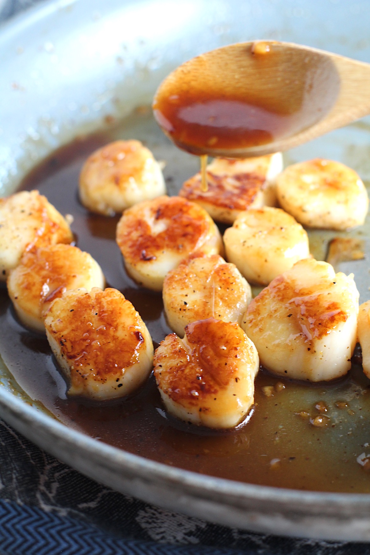 Wood spoon drizzling glaze on Glazed Pan Seared Scallops with Garlic and Honey in a pan with spatula.
