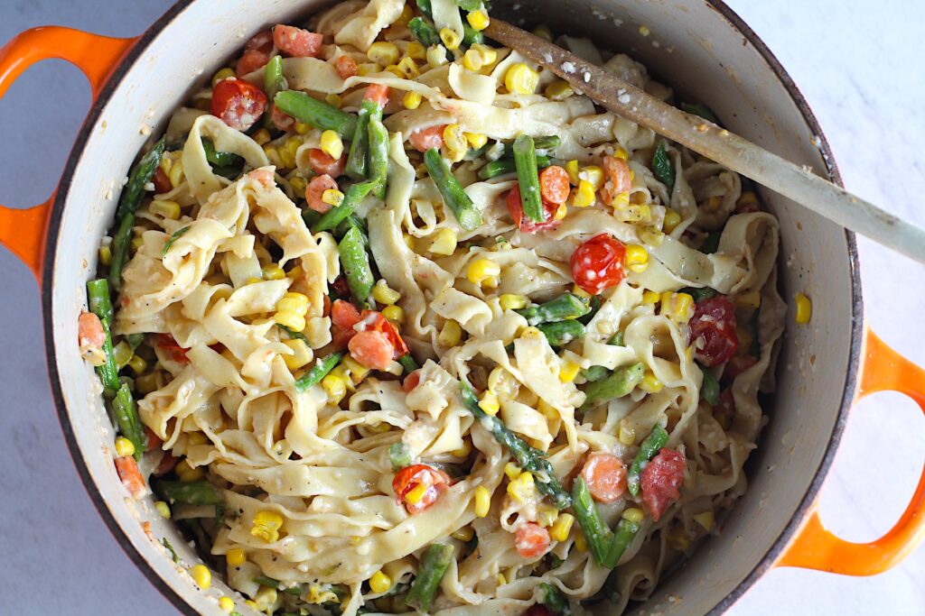 Fresh, Creamy Pasta Primavera Recipe in a pot on counter with fettuccine, parmesan cheese, roasted asparagus, carrots, tomatoes and fresh corn kernels.