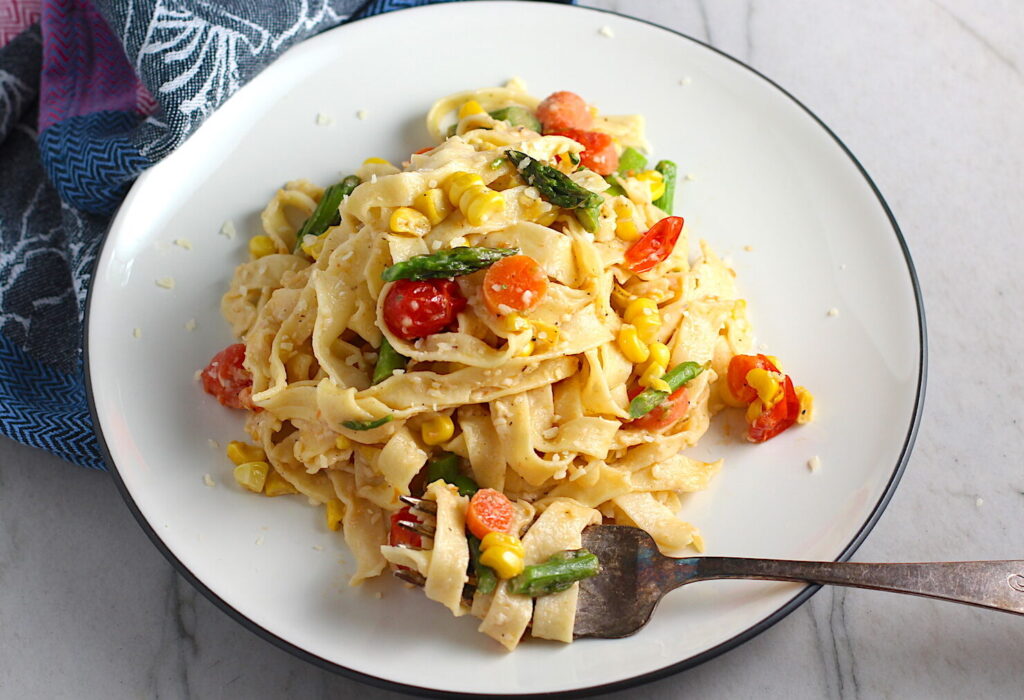Fresh, Creamy Pasta Primavera Recipe on a plate with fork on counter. It has fettuccine, parmesan cheese, roasted asparagus, carrots, tomatoes and fresh corn kernels.