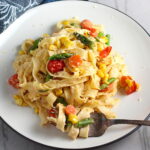 Fresh, Creamy Pasta Primavera Recipe on a plate with fork on counter. It has fettuccine, parmesan cheese, roasted asparagus, carrots, tomatoes and fresh corn kernels.