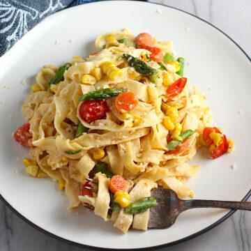 Fresh, Creamy Pasta Primavera Recipe on a plate with fork on counter. It has fettuccine, parmesan cheese, roasted asparagus, carrots, tomatoes and fresh corn kernels.