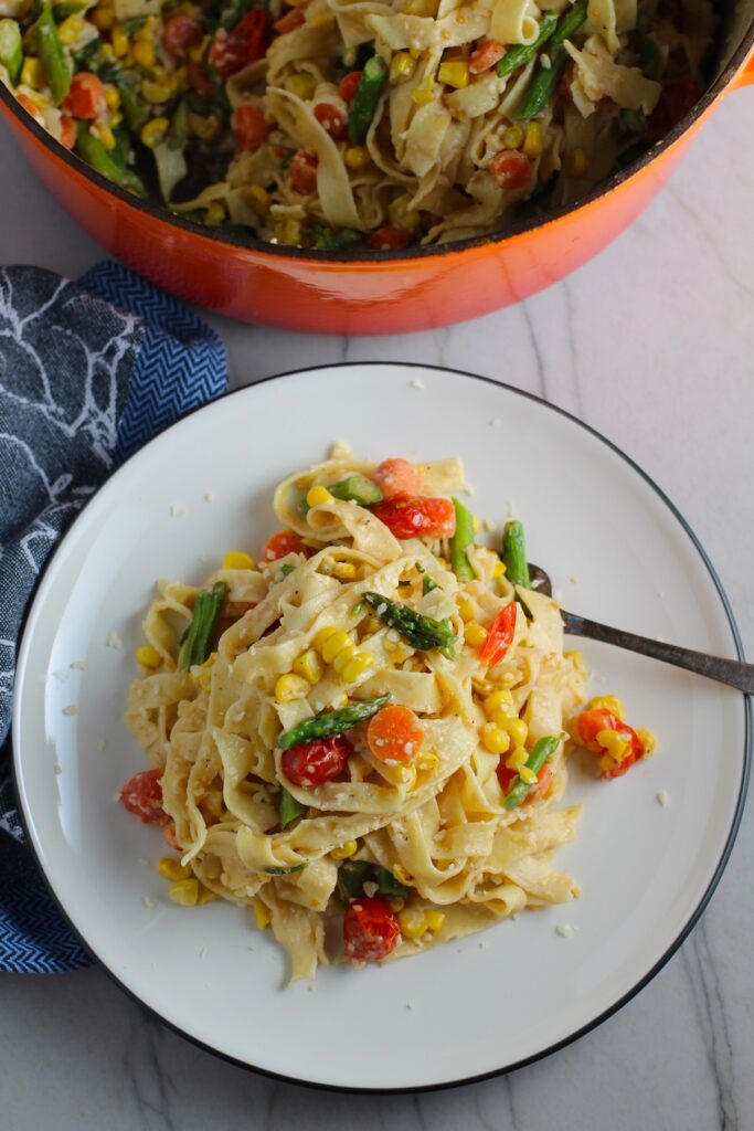 Fresh, Creamy Pasta Primavera Recipe on a plate with fork and pot in background on counter. It has fettuccine, parmesan cheese, roasted asparagus, carrots, tomatoes and fresh corn kernels.