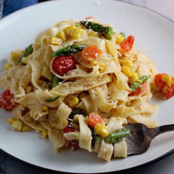 Fresh, Creamy Pasta Primavera Recipe on a plate with fork on counter. It has fettuccine, parmesan cheese, roasted asparagus, carrots, tomatoes and fresh corn kernels.