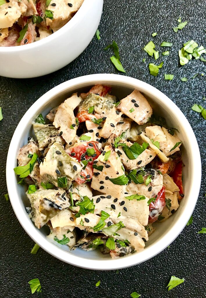 Grilled Teriyaki Chicken Salad in a bowl on black background with chicken, green peppers, tomatoes, onions, black sesame seeds, and cilantro.