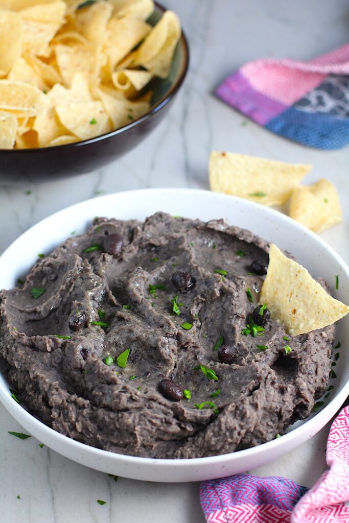Creamy, rich, smokey, and delicious refried black beans recipe in a bowl with a tortilla chip dipped in and a few black beans and chopped cilantro sprinkled on top. On the counter are tortilla chips in a bowl.