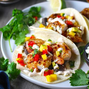 Grilled Shrimp Tacos in corn tortillas on a plate with Mango Salsa & Chipotle Crema. On the side are bowls of cotija cheese, crema, salsa, and cilantro leaves.