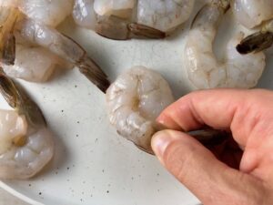 Hand pinching off the tail from a raw shrimp with more shrimp on the plate for Grilled Shrimp Tacos