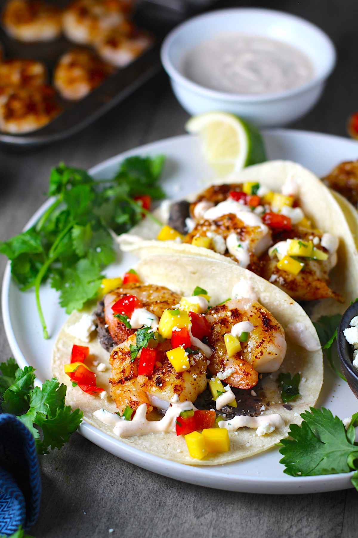 Grilled Shrimp Tacos in corn tortillas on a plate with Mango Salsa & Chipotle Crema. On the side are bowls of cotija cheese, crema, salsa, and cilantro leaves.