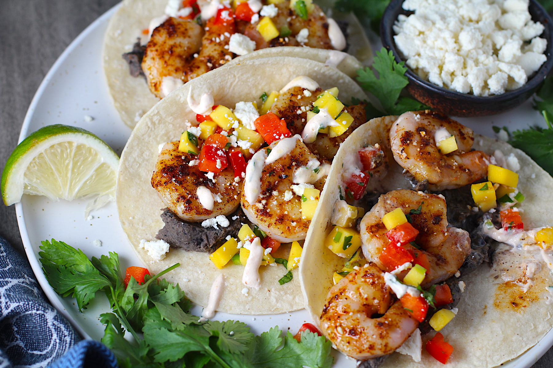 Grilled Shrimp Tacos in corn tortillas on a plate with Mango Salsa & Chipotle Crema. On the side are bowls of cotija cheese, crema, salsa, and cilantro leaves.